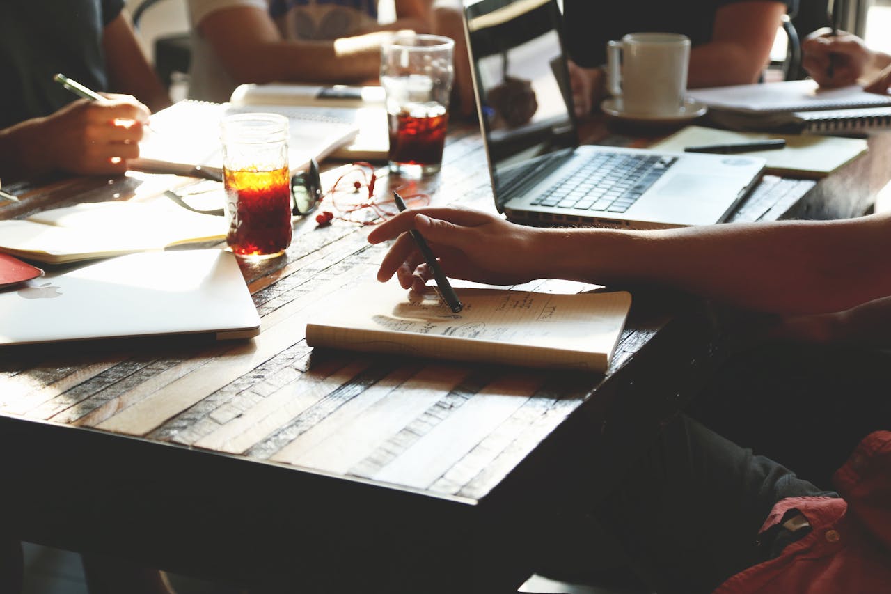 A group of adults engaged in a collaborative office meeting, discussing ideas with notes, laptops, and refreshments.